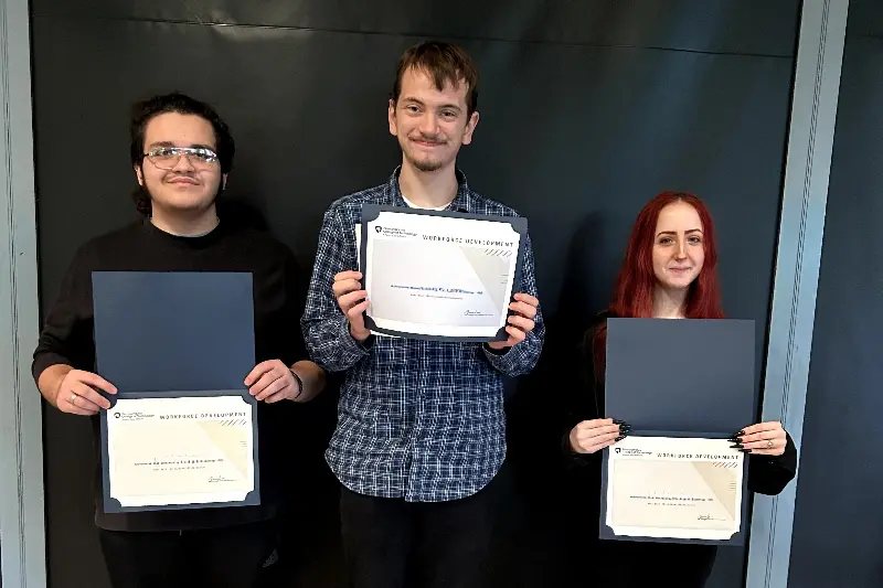 Three Insight PA students are standing against a black wall holding up their Workforce Development Certificates.