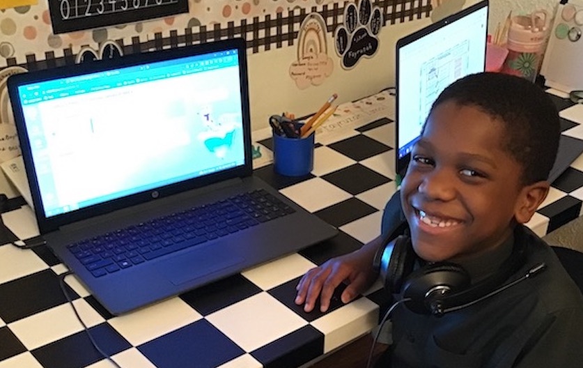 An Insight PA student is sitting in front of his laptop and smiling at the camera. He is sitting at a checkerboard desk in his home learning space with classroom decorations on the wall.