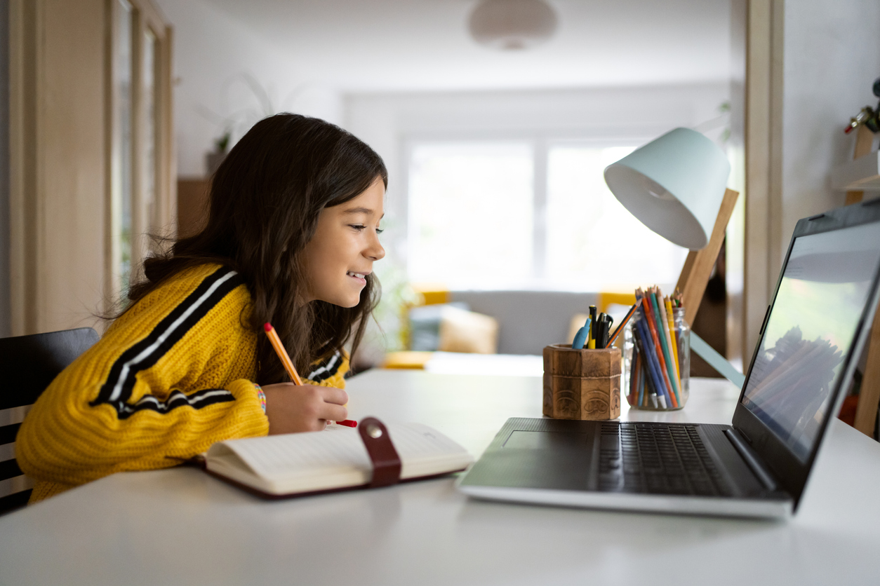 Stock image of a young student in front of their laptop. They are sitting at a white table with a lamp and jars of pencils and writing in a notebook.