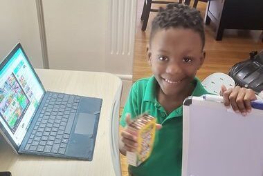 A young student proudly holds up their schoolwork while sitting in front of a lap top