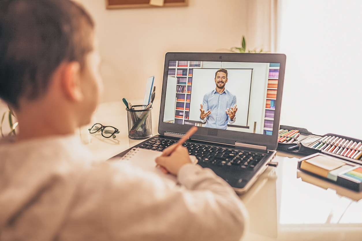 A student is sitting at their desk in front of a laptop screen. On the screen is a teacher who is working with the student one-on-one.