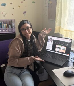 The image shows a young woman sitting at a desk, smiling and gesturing with her hands. Behind her, the wall is decorated with various stickers and artwork.